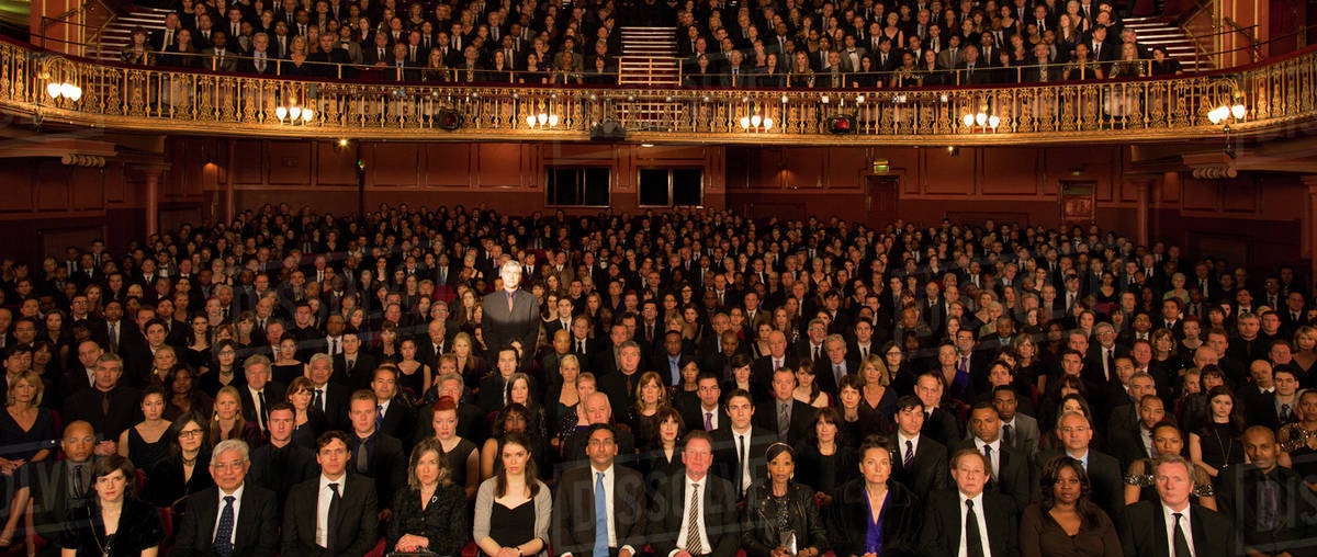 A packed theatre auditorium of seated audience members facing the camera