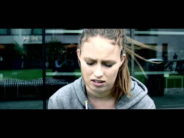Still from the Unlockhiddenweb video: a young woman in a grey hooded top, hair blown across her face, looking down, in front of a glass building